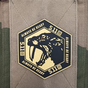 May include: A black and gold patch with the text "ALWAYS BE READY" and "5.11". The patch features a saber-toothed tiger design. The patch is on a tan fabric background with a camouflage pattern.