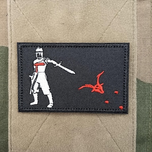 May include: A rectangular black embroidered patch featuring a white knight figure with a red cross on the chest, holding a sword pointed towards a red dragon skull graphic. The patch is stitched onto a tan fabric square with a camouflage background.