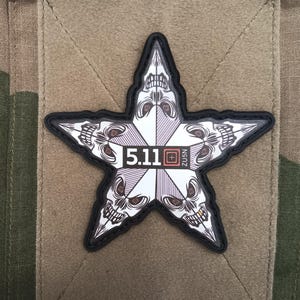 Peut inclure: Écusson en forme d'étoile avec un motif de crâne et le texte "5.11". L'écusson a un bord noir et est posé sur un carré de tissu beige, sur un fond de camouflage. L'étoile a un design blanc et violet.