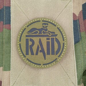 Peut inclure: Écusson rond vert olive avec le mot "RAID" en gris foncé et une figure de sphinx. Le patch est cousu sur un carré de tissu beige, sur un fond de camouflage. Thème militaire ou tactique.