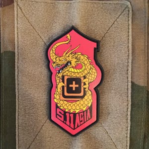 Peut inclure: Un écusson rouge et or avec un motif de dragon doré et le texte "SIT ASIA" sous un symbole de croix blanche. L'écusson est posé sur un fond beige et un motif de camouflage.