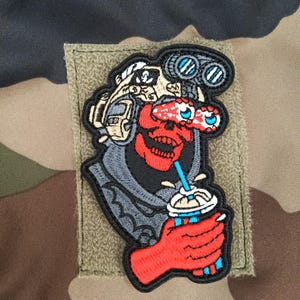Peut inclure: Écusson brodé représentant un crâne rouge portant un casque et des jumelles, tenant une boisson avec une paille. L'écusson a une bordure noire et est posé sur un fond de camouflage.