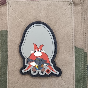 Puede incluir: Un parche de dibujos animados con un personaje con barba roja y un arma. El parche está sobre un cuadrado beige con un fondo de camuflaje. El personaje tiene una cara gris y lleva pantalones azules.