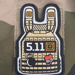 May include: A tan and black patch in the shape of a tactical vest. The patch features the text "5.11" and a plus sign, along with a bomb and a heart with the word "PALIO". The patch is on a camouflage background.