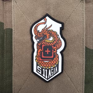 May include: A patch with an orange dragon design, a red cross symbol, and the text "S.11 ASIA" on a white and black background. The patch is set against a tan rectangular patch on a camouflage fabric.