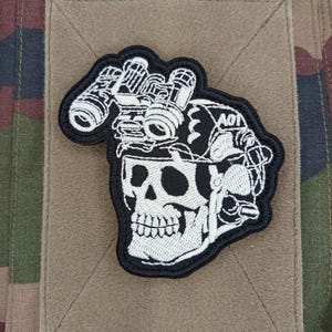 Peut inclure: Écusson brodé noir et blanc représentant un crâne portant un casque avec des lunettes de vision nocturne. L'écusson est posé sur un carré de tissu beige, sur un fond de camouflage.