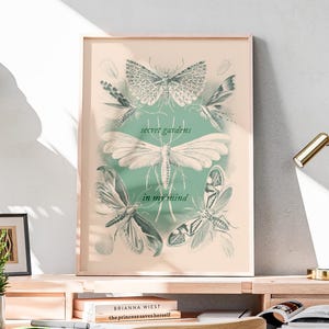 May include: A light wood desk with a framed print featuring a butterfly design and the text "in the secret garden". A small potted plant, books, and a gold desk lamp are on the desk. A wooden chair is in front of the desk.