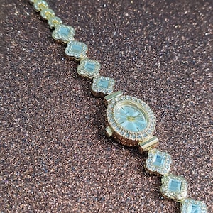 Puede incluir: Un reloj dorado con una esfera blanca y una pulsera engastada con cristales. La pulsera est&aacute; hecha de peque&ntilde;os cristales de forma cuadrada.