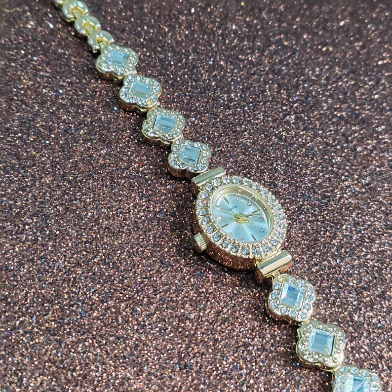 Puede incluir: Un reloj dorado con una esfera blanca y una pulsera engastada con cristales. La pulsera est&aacute; hecha de peque&ntilde;os cristales de forma cuadrada.