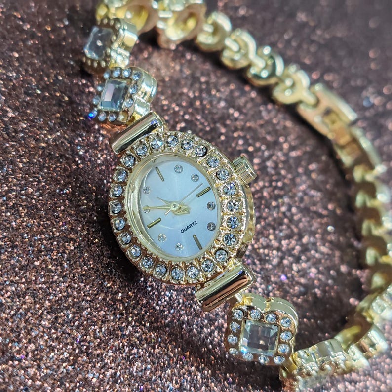 Puede incluir: Un reloj dorado con una esfera blanca y una pulsera engastada con cristales. La esfera del reloj tiene un peque&ntilde;o marcador en forma de diamante en la posici&oacute;n de las 12 en punto y la palabra "QUARTZ" impresa debajo del centro de la esfera.