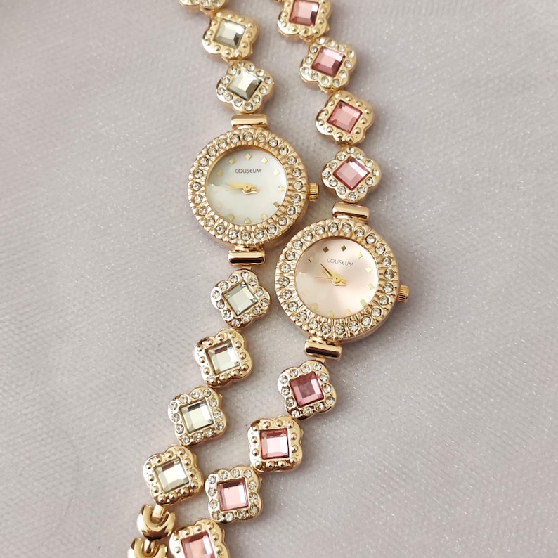 Puede incluir: Dos relojes de tono dorado con biseles y correas engastados con cristales. Las esferas de los relojes son blancas y rosadas con la palabra "COLISEUM" impresa en ellas.