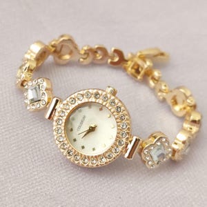 Puede incluir: Un reloj dorado con una esfera blanca y una pulsera engastada con cristales. La esfera del reloj tiene la palabra "COUSEUM" impresa.