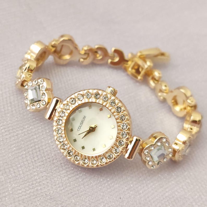 Puede incluir: Un reloj dorado con una esfera blanca y una pulsera engastada con cristales. La esfera del reloj tiene la palabra "COUSEUM" impresa.