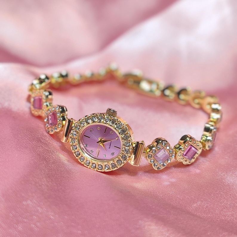 Puede incluir: Un reloj dorado con una esfera rosa y una pulsera engastada con cristales. La pulsera presenta piedras rosas y transparentes.