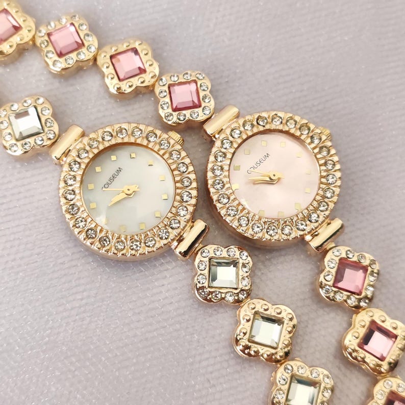 Puede incluir: Dos relojes de tono dorado con correas y car&aacute;tulas engastadas con cristales. Las car&aacute;tulas de los relojes son blancas y rosadas con la palabra "OLEGSUM" impresa en ellas.