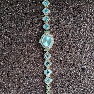 Puede incluir: Un reloj de pulsera de tono dorado con una esfera blanca y una pulsera engastada con cristales. La pulsera est&aacute; compuesta por peque&ntilde;os eslabones de forma cuadrada.