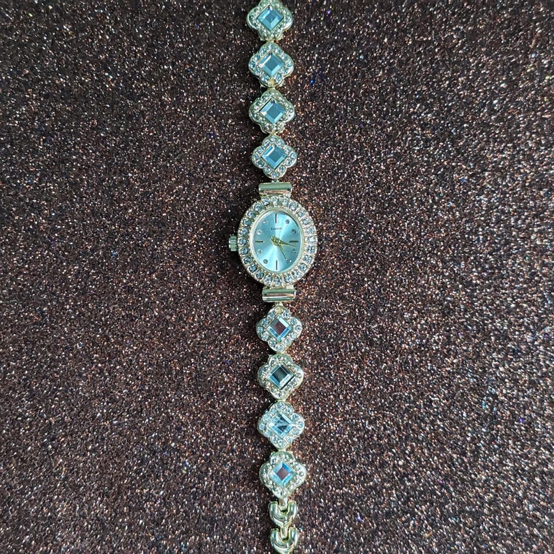 Puede incluir: Un reloj de pulsera de tono dorado con una esfera blanca y una pulsera engastada con cristales. La pulsera est&aacute; compuesta por peque&ntilde;os eslabones de forma cuadrada.