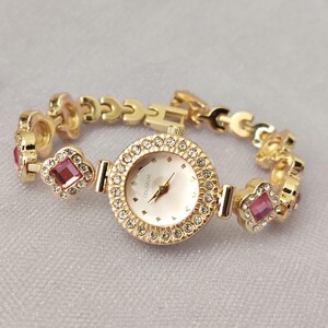 Puede incluir: Un reloj dorado con una esfera rosa y una pulsera engastada con cristales. La esfera del reloj tiene la palabra "COUSSEUM" impresa.