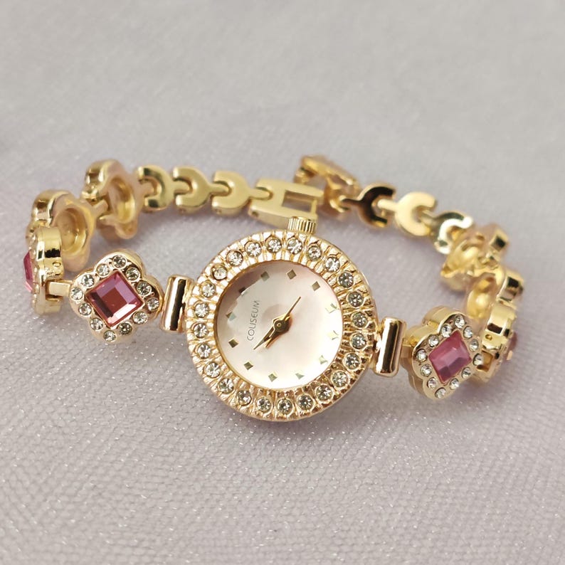 Puede incluir: Un reloj dorado con una esfera rosa y una pulsera engastada con cristales. La esfera del reloj tiene la palabra "COUSSEUM" impresa.