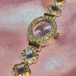 Puede incluir: Un reloj dorado con una esfera rosa y una pulsera engastada con cristales. La pulsera est&aacute; formada por una serie de formas de tr&eacute;bol de cuatro hojas unidas, cada una con un cristal rosa en el centro.