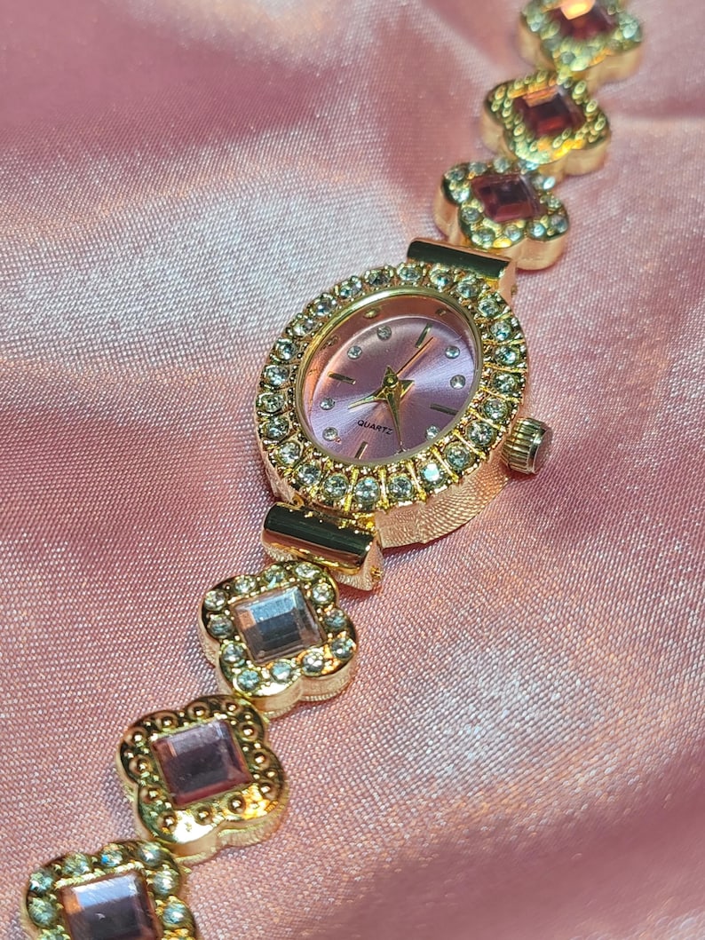 Puede incluir: Un reloj dorado con una esfera rosa y una pulsera engastada con cristales. La pulsera est&aacute; formada por una serie de formas de tr&eacute;bol de cuatro hojas unidas, cada una con un cristal rosa en el centro.