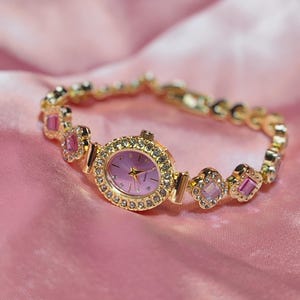 Puede incluir: Un reloj dorado con una esfera rosa y una pulsera engastada con cristales. La pulsera presenta piedras rosas y transparentes.