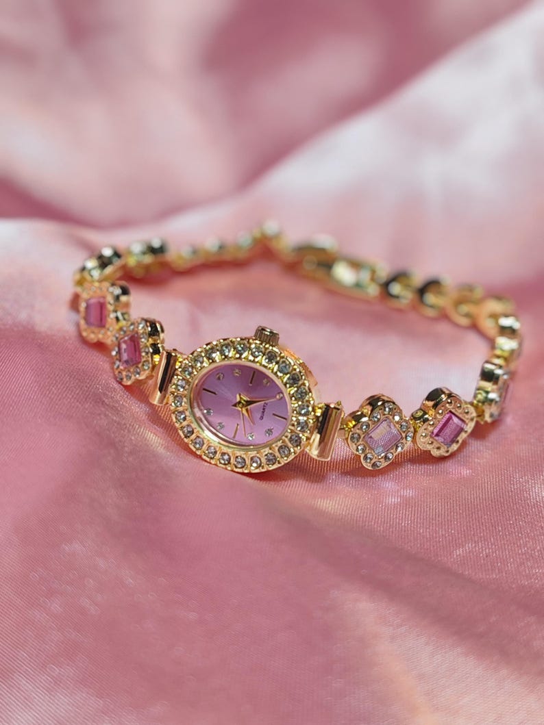 Puede incluir: Un reloj dorado con una esfera rosa y una pulsera engastada con cristales. La pulsera presenta piedras rosas y transparentes.