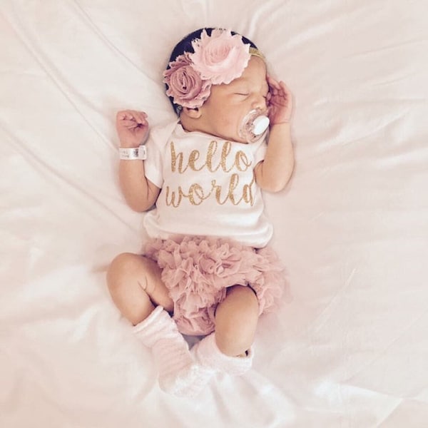 May include: A baby girl wearing a white onesie with gold glitter lettering that says "hello world". She is also wearing a pink tutu and a pink flower headband. 