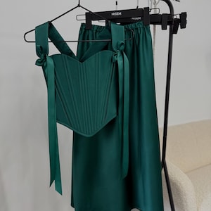 May include: A two-piece emerald green outfit, consisting of a corset top and a midi skirt, hangs on a black clothing rack. The corset features vertical seams and tie straps. The skirt has a flowing silhouette. The outfit is displayed in a store setting.