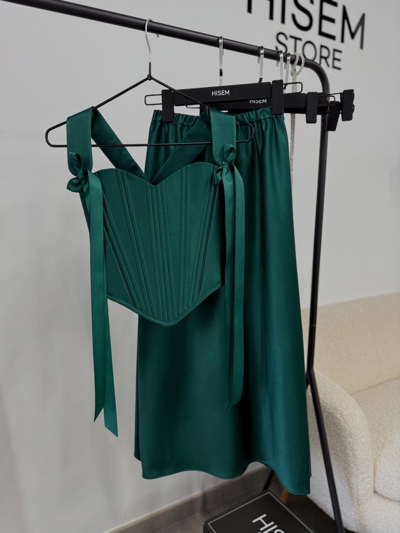 May include: A two-piece emerald green outfit, consisting of a corset top and a midi skirt, hangs on a black clothing rack. The corset features vertical seams and tie straps. The skirt has a flowing silhouette. The outfit is displayed in a store setting.