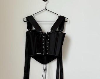 May include: Black corset top with adjustable straps and a lace-up front. The top features a structured bodice and long, flowing ribbons. The word "HISEM" is printed on the front. The corset is hanging on a black hanger against a white wall.