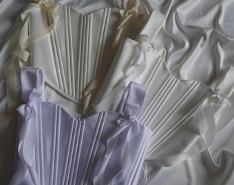 May include: Several white satin corsets with vertical boning and ribbon straps are arranged on a white satin surface. The corsets have a classic silhouette, with a sweetheart neckline and a fitted waist. The ribbons are tied into bows.