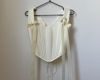 May include: Ivory-colored corset top with vertical ribbing and cream-colored ribbon straps tied into bows. The corset has a V-neckline and a lace-up back. The top is hanging on a black hanger against a white wall.