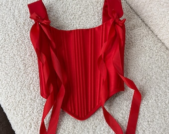 May include: A red satin corset with boning and adjustable straps. The corset has long red ribbon ties that can be tied in the front or back.