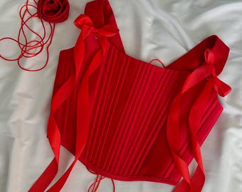 May include: A red satin corset with a bustier style and two long red satin ribbons tied into bows at the shoulders. The corset has a lace-up back and is made of a pleated fabric.