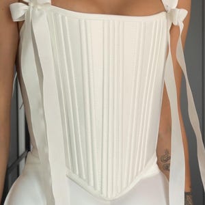 May include: A white corset top with vertical stitching and ribbon straps tied into bows. The top is part of a dress, with a flowing skirt visible below. The overall style is elegant and suitable for special occasions.