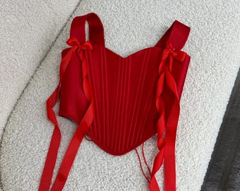 May include: A red satin corset with a sweetheart neckline and two bows on the straps. The corset has a boned structure and is designed to cinch the waist.