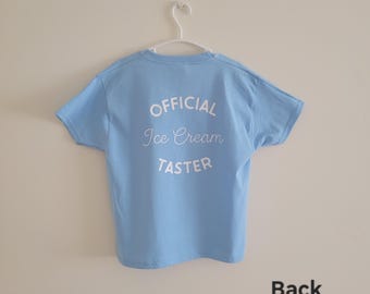 May include: A light blue t-shirt with the words "OFFICIAL Ice Cream TASTER" printed in white on the back. The shirt is hanging on a white hanger against a plain wall. The word "Back" is printed in black at the bottom.
