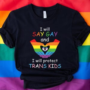以下が含まれることがあります： 「I will SAY GAY and I will protect TRANS KIDS」とカラフルな文字で書かれた黒いTシャツ。デザインには、虹色の手でトランスジェンダーの旗の色でハートを抱えています。Tシャツは虹色の旗の背景に表示されています。