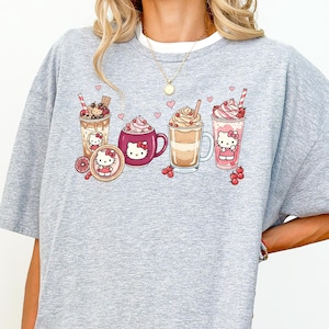 May include: A grey t-shirt with a graphic of cartoon beverages, including coffee, milkshake, and hot chocolate. The design features a cartoon character. The shirt has a white collar and a loose fit.
