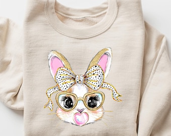 May include: A beige sweatshirt with a graphic of a bunny wearing pink and black polka dot bow and gold-rimmed glasses. The bunny is holding a pink heart.