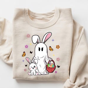 Puede incluir: Una sudadera beige con un conejito fantasma blanco que sostiene una cesta de huevos de Pascua y un conejito blanco más pequeño a su lado. El diseño está rodeado de mariposas y flores.