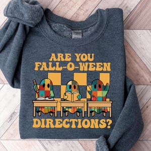 Könnte beinhalten: Ein dunkelgraues Sweatshirt mit dem Text "ARE YOU FALL-O-WEEN DIRECTIONS?" in Orange. Die Grafik zeigt drei geisterhafte Figuren an Schreibtischen mit Patchwork-Designs. Der Hintergrund ist eine Holzoberfläche.