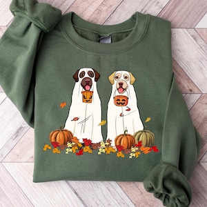 May include: A green sweatshirt with a graphic of two dogs dressed as ghosts for Halloween. The dogs are holding jack-o'-lanterns and surrounded by fall leaves.