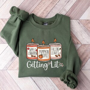 May include: A green sweatshirt with a graphic of three candles with the text "Apple Cinnamon", "Pumpkin Spice", and "Falling Leaves" on them. The text "Getting Lit" is below the candles.