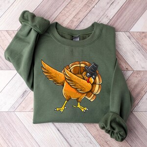 Può includere: Una felpa verde con un'immagine di un tacchino dei cartoni animati che indossa occhiali da sole e un cappello, con le ali spiegate in una posa 'dab'.