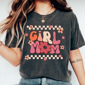 May include: A dark grey t-shirt with a retro-style graphic that says "Girl Mom" in pink, orange, and yellow letters with flowers around the text. The graphic is on a checkered background.