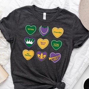 Peut inclure: Un t-shirt gris fonc&eacute; avec un motif de neuf c&oelig;urs violets, verts et jaunes. Chaque c&oelig;ur a un design et un texte diff&eacute;rents, notamment "Bends", "King Cake", "Mardi Gras" et "Fat Tuesday".