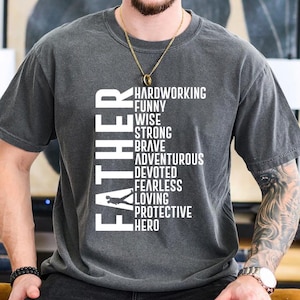 Op de afbeelding: Een donkergrijs T-shirt met een witte afbeelding die verticaal "Father" zegt met de woorden "Hardworking, Funny, Wise, Strong, Brave, Adventurous, Devoted, Fearless, Loving, Protective, Hero" die verticaal ernaast staan.