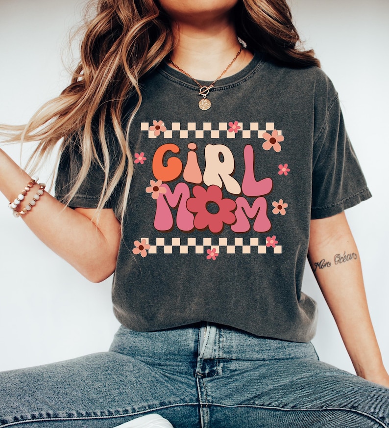 May include: A dark grey t-shirt with a retro-style graphic that says "Girl Mom" in pink, orange, and yellow letters with flowers around the text. The graphic is on a checkered background.
